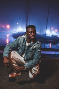 Young man looking away while crouching at harbor in night