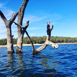 Teenage boy standing on bare tree over lake against blue sky