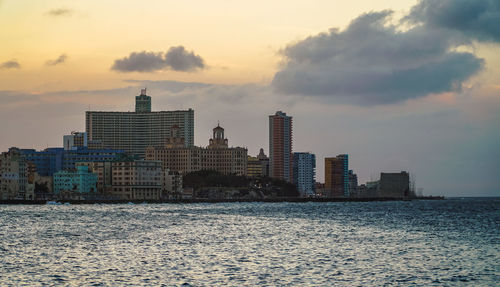 Sea by buildings in city against sky during sunset