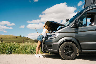 Side view of unrecognizable traveling female looking under opened hood of camper van while having problems during trip through summer nature