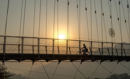 Silhouette of woman riding bicycle on footbridge against sky during sunset