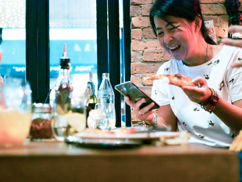 Cheerful woman using phone while having pizza at table in restaurant