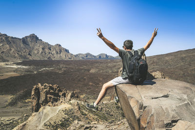 Rear view of man sitting on rock with arms raised against clear sky