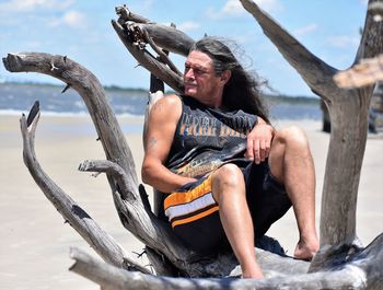 Man sitting on driftwood at beach against sky