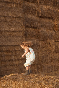 Side view of carefree child in overalls jumping on straw bale on sunny day in countryside