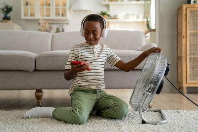 Happy african american child boy relaxing with smartphone in front of electric fan at home
