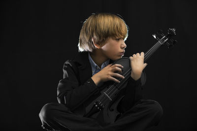 Boy holding violin while sitting against black background