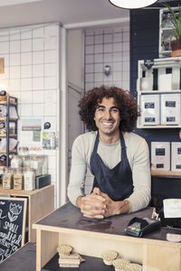 Happy male owner with hands clasped leaning on checkout counter at organic shop