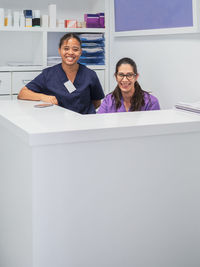 Female nurses in scrubs smiling while working behind counter on reception of modern clinic looking at camera