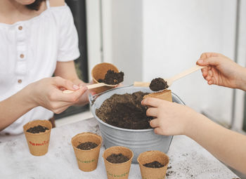 Two girls pour earth with wooden spoons from a zinc bucket into cardboard cups.