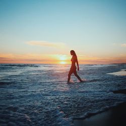 Woman wearing bikini while walking in sea against sky during sunset