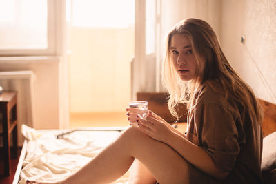 Young woman holding glass of water while sitting in bed at home