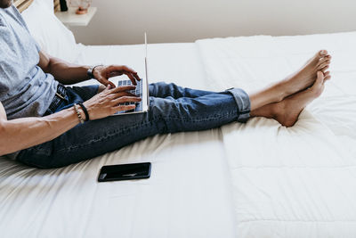 Young man working with laptop and mobile phone while sitting on bed at home