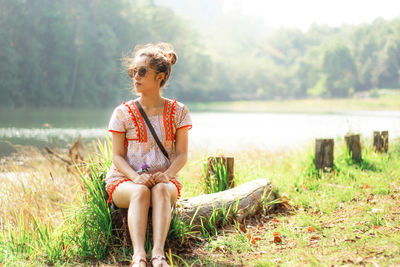 Young woman sitting on grass by lake against sky
