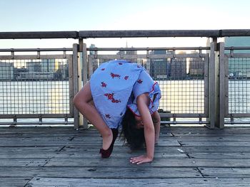 Side view of girl bending backwards on pier by river against clear sky in city during sunset