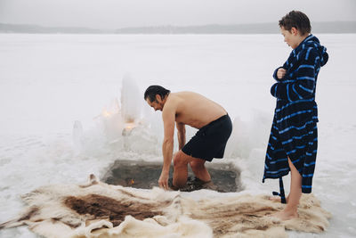 Boy looking at father taking ice bath at frozen lake