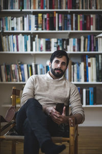 Portrait of young man holding smart phone while sitting against bookshelf