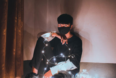 Portrait of young man with illuminated string lights while sitting against wall
