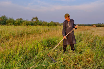 An senior muslim man in a skullcap and traditional clothes mows hand-scythe grass in a hayfield. 