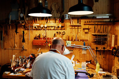 Back view of unrecognizable male luthier master sitting on chair and catching tool from wall while making instruments in workshop