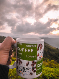 Hand holding coffee cup against sky at sunset