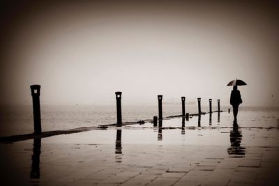 Silhouette man standing on wooden post by sea against sky during rainy season