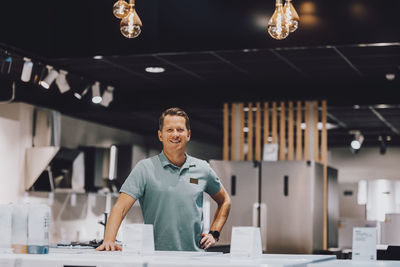 Portrait of confident male retail clerk standing with hand on hip in electronics store