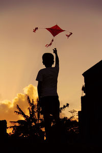 Rear view of silhouette boy flying kite against sky during sunset