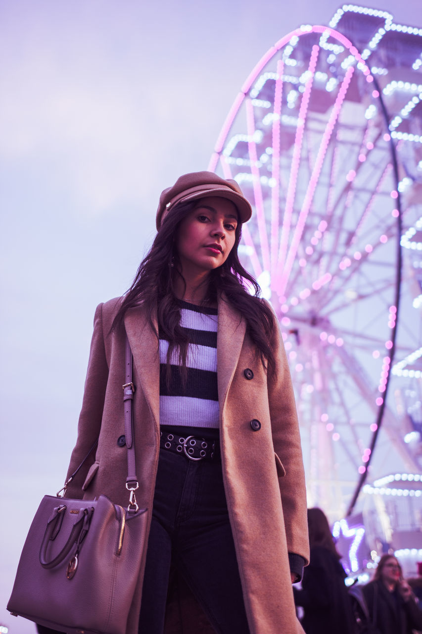 clothing, real people, leisure activity, lifestyles, amusement park, amusement park ride, ferris wheel, front view, arts culture and entertainment, portrait, one person, hat, illuminated, standing, three quarter length, sky, women, young adult, low angle view, outdoors, beautiful woman, hairstyle, warm clothing