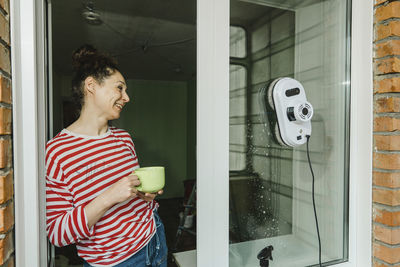 Happy woman with coffee cup cleaning window through robot washer