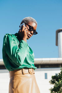 Confident young african american businesswoman with short dyed hair in stylish clothes and sunglasses talking on mobile phone while standing on street against cloudless blue sky