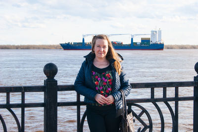 Portrait of smiling young woman standing by railing against river