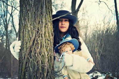 Portrait of teenage girl with doll standing by tree during winter