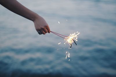 Cropped hand of woman holding illuminated sparklers against sea during sunset