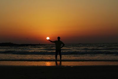 Optical illusion on silhouette man holding sun while standing beach against orange sky