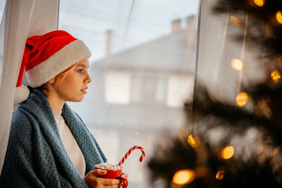 Young teen girl in santa hat having a cup of hot chocolate with marshmallow with christmas tree on