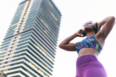 Low angle of young fit female in sportswear and headphones listening to music while standing on street against modern building