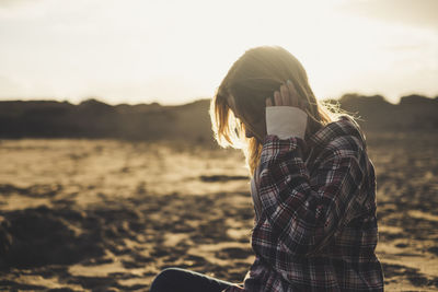 Side view of woman sitting at beach against sky during sunset