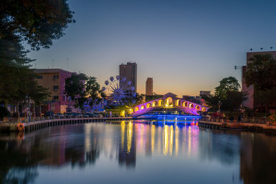 Illuminated bridge over river by buildings against sky at dusk