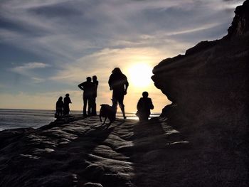 Silhouette people with dog standing on cliff at shore