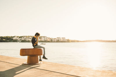 Side view of teenage boy answering mobile phone while sitting on bollard by river against clear sky during sunny day