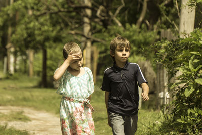 Siblings walking on field against trees on sunny day