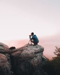 Side view of hiker photographing on cliff against sky during sunset