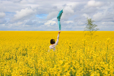 Rear view of woman throwing scarf in mid-air while standing amidst rapeseed field against cloudy sky