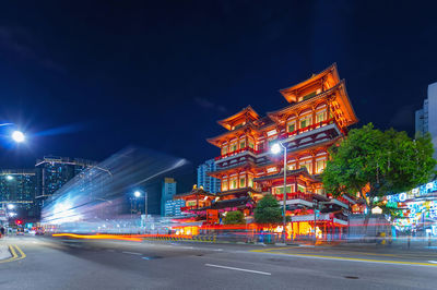 Illuminated city street by buildings against sky at night