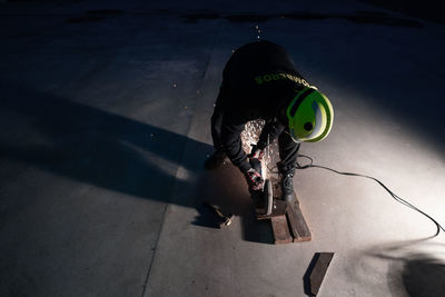 From above full body of unrecognizable male welder in protective helmet and goggles cutting iron details with chop saw during work at industrial factory at night