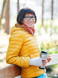 Happy wide smiling women in bright yellow jacket is holding thermos mug. hot tea or other beverage