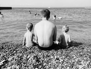 Rear view of father and sons sitting on sea shore against sky