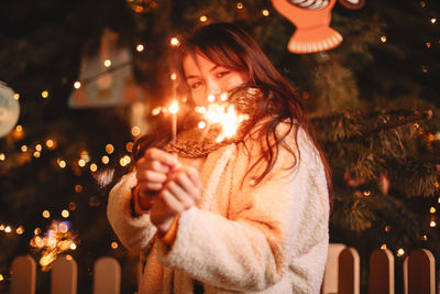 Teenage girl holding sparklers standing by christmas tree at night