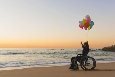 People sitting on balloons at beach against sky during sunset